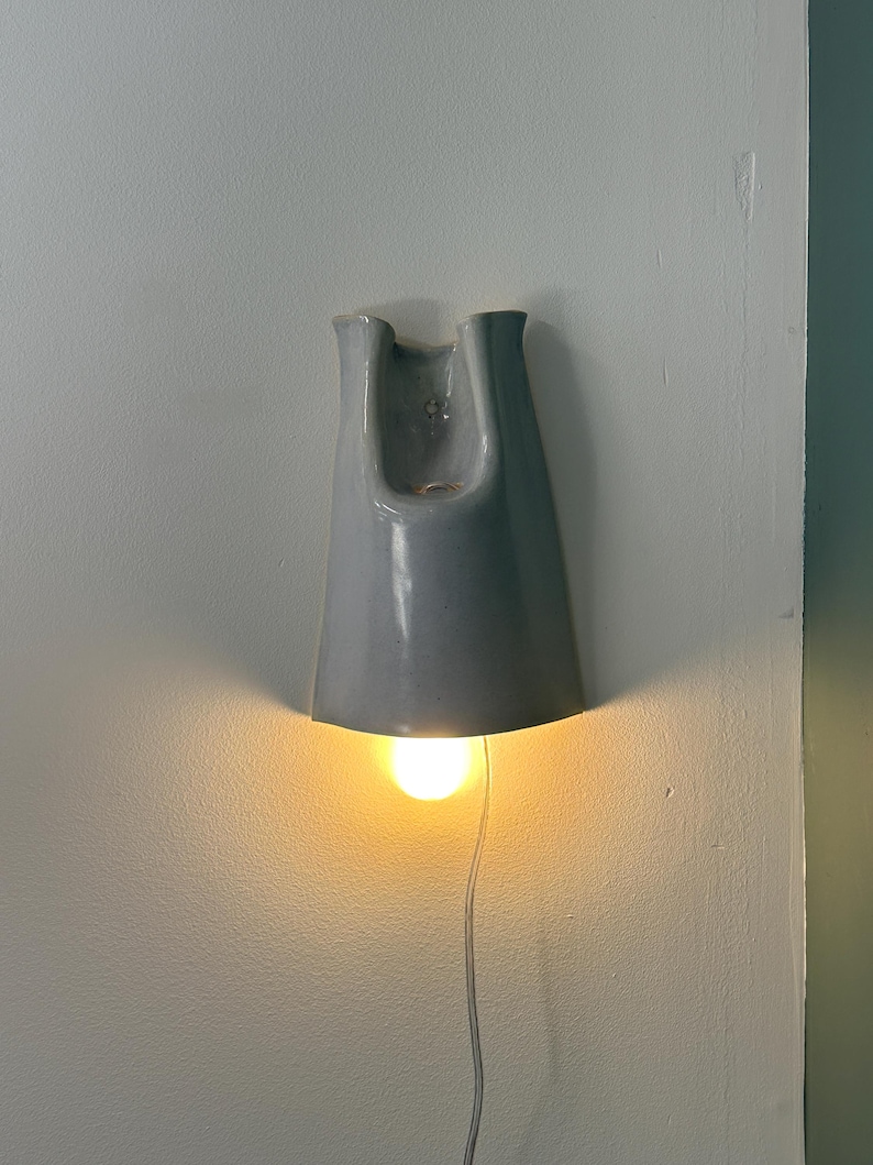 K&ouml;nnte beinhalten: Eine Wandleuchte aus Keramik mit einer glatten, gl&auml;nzenden hellgrauen Oberfl&auml;che. Die Leuchte hat eine ausgestellte, glockenf&ouml;rmige Gestalt mit einer offenen Oberseite und einer nach unten gerichteten &Ouml;ffnung, die warmes Licht abgibt.