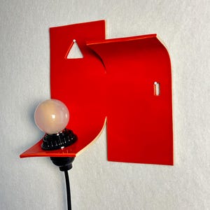 May include: A contemporary wall light with a vibrant red geometric shape and a small shelf. A spherical light bulb sits on a black base, connected to a black cable. The design is minimalist and artistic, suitable for modern interiors.