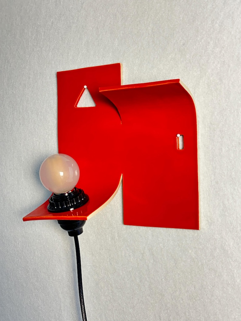 May include: A contemporary wall light with a vibrant red geometric shape and a small shelf. A spherical light bulb sits on a black base, connected to a black cable. The design is minimalist and artistic, suitable for modern interiors.