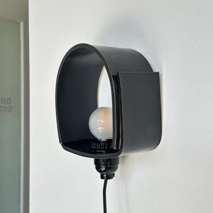 Puede incluir: Una l&aacute;mpara de pared negra moderna con un dise&ntilde;o curvo y rectangular. Incluye una bombilla y un cable el&eacute;ctrico negro. La l&aacute;mpara est&aacute; montada en una pared blanca, ofreciendo una est&eacute;tica minimalista.