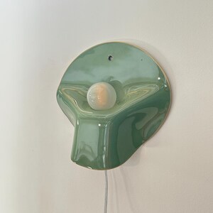 May include: A wall-mounted light fixture featuring a glossy, sage green ceramic shade with an organic, asymmetrical shape. A single round, frosted white light bulb is centered on the surface, and a thin white power cord hangs from the bottom.