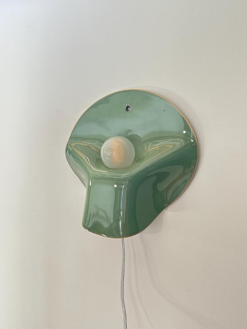 May include: A wall-mounted light fixture featuring a glossy, sage green ceramic shade with an organic, asymmetrical shape. A single round, frosted white light bulb is centered on the surface, and a thin white power cord hangs from the bottom.