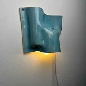 May include: A light blue ceramic wall sconce with a fluid, organic shape and a glossy finish. The fixture features two small circular mounting holes at the top corners and emits a warm, glowing light from a single bulb positioned underneath.