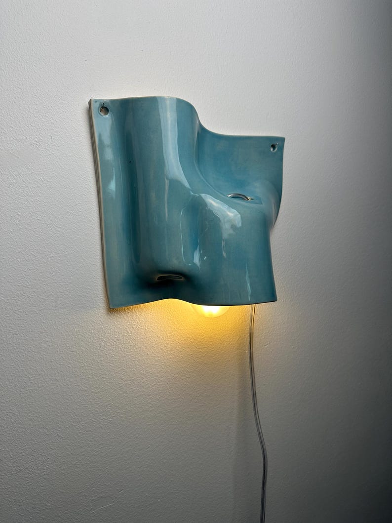 May include: A light blue ceramic wall sconce with a fluid, organic shape and a glossy finish. The fixture features two small circular mounting holes at the top corners and emits a warm, glowing light from a single bulb positioned underneath.