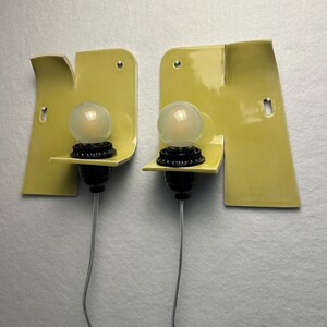May include: A pair of wall-mounted light fixtures featuring asymmetrical, pale yellow ceramic backplates with a glossy finish. Each fixture includes a black socket holding a round, frosted glass light bulb and a thin, clear electrical cord.