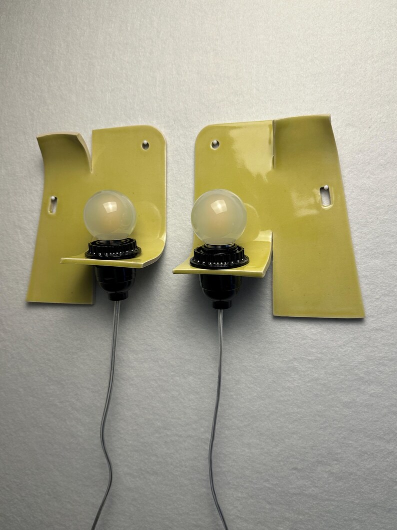 May include: A pair of wall-mounted light fixtures featuring asymmetrical, pale yellow ceramic backplates with a glossy finish. Each fixture includes a black socket holding a round, frosted glass light bulb and a thin, clear electrical cord.