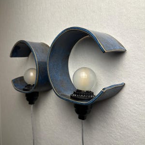 May include: Two wall-mounted sconces featuring a curved, C-shaped blue ceramic shade with a dark, mottled finish. Each fixture includes a round, frosted white light bulb seated in a black socket, with a clear power cord extending downward.