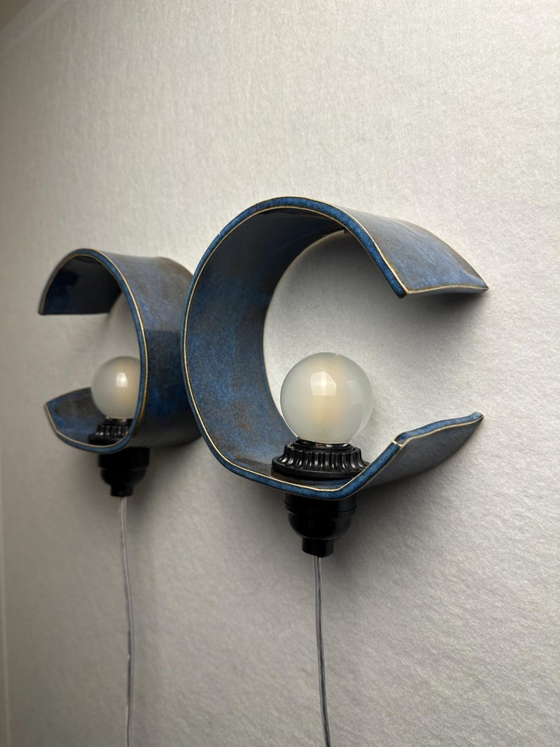 May include: Two wall-mounted sconces featuring a curved, C-shaped blue ceramic shade with a dark, mottled finish. Each fixture includes a round, frosted white light bulb seated in a black socket, with a clear power cord extending downward.