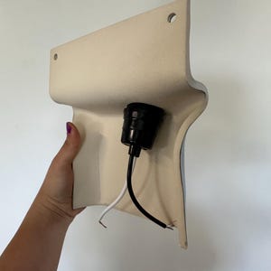 Peut inclure: Une base de luminaire murale incurv&eacute;e de couleur cr&egrave;me dot&eacute;e d'une douille centrale noire. Deux petits trous circulaires sont situ&eacute;s en haut pour la fixation. Un fil &eacute;lectrique noir et un blanc sortent de la douille pour le raccordement.
