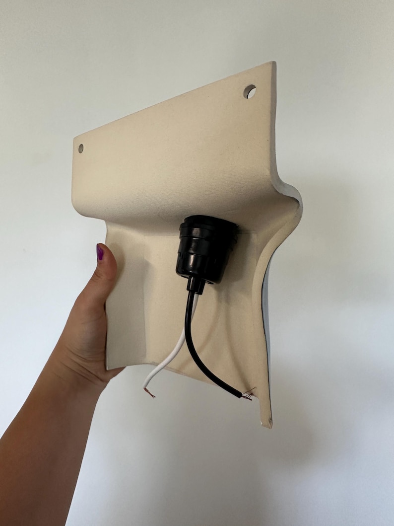 Peut inclure: Une base de luminaire murale incurv&eacute;e de couleur cr&egrave;me dot&eacute;e d'une douille centrale noire. Deux petits trous circulaires sont situ&eacute;s en haut pour la fixation. Un fil &eacute;lectrique noir et un blanc sortent de la douille pour le raccordement.