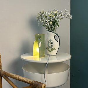May include: A light green ceramic lamp with a curved, open-sided structure emitting a warm glow, positioned next to a white ceramic pitcher featuring a green handle and a painted floral design filled with white and yellow baby's breath flowers.