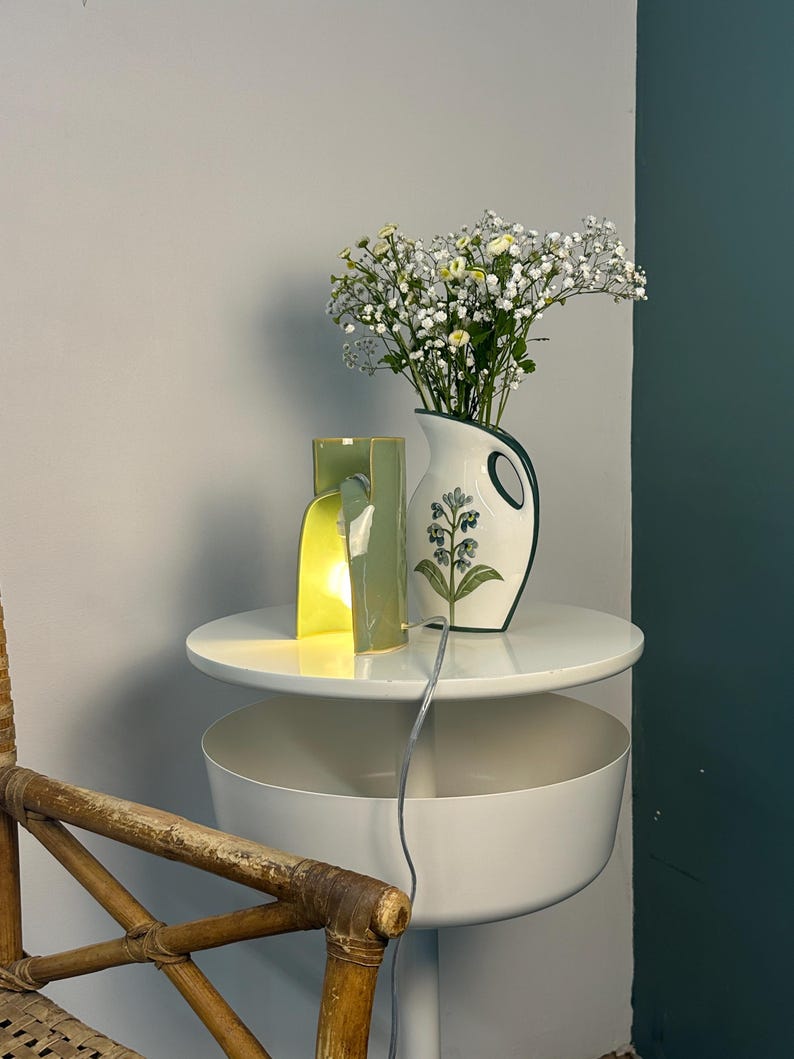 May include: A light green ceramic lamp with a curved, open-sided structure emitting a warm glow, positioned next to a white ceramic pitcher featuring a green handle and a painted floral design filled with white and yellow baby's breath flowers.