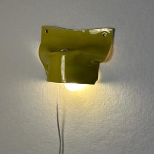 May include: A unique, olive-green wall sconce with a textured, folded design. The sconce has a cylindrical base and a bright light bulb. A thin, silver wire extends from the base.