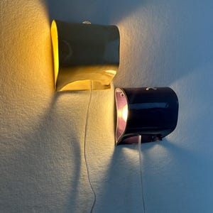 May include: Two wall-mounted lamps with curved, glossy ceramic shades. One lamp is yellow and emits a warm glow, while the other is dark purple with a visible light bulb inside. Both fixtures feature thin, clear power cords hanging downwards.