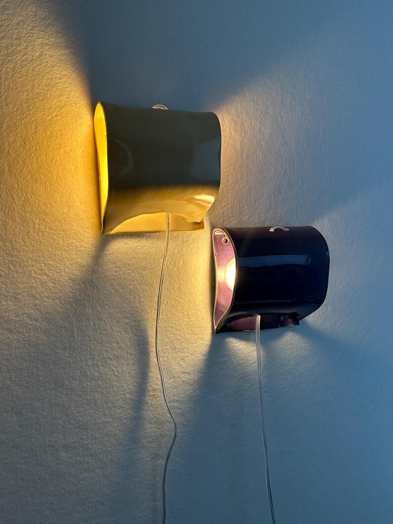 May include: Two wall-mounted lamps with curved, glossy ceramic shades. One lamp is yellow and emits a warm glow, while the other is dark purple with a visible light bulb inside. Both fixtures feature thin, clear power cords hanging downwards.