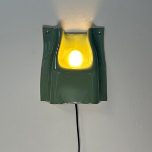 May include: A wall-mounted ceramic sconce featuring a glossy sage green exterior and a bright yellow interior. The fixture has a curved, folded shape with a central opening that emits a warm, glowing light. A thin black power cord hangs downwards.