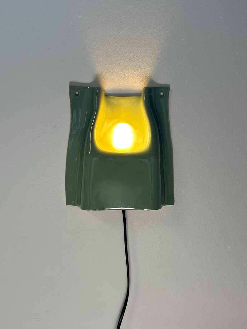 May include: A wall-mounted ceramic sconce featuring a glossy sage green exterior and a bright yellow interior. The fixture has a curved, folded shape with a central opening that emits a warm, glowing light. A thin black power cord hangs downwards.