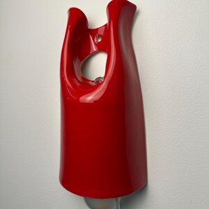 May include: A glossy red wall-mounted light fixture with a smooth, curved, organic shape. The fixture features a central circular cutout near the top for mounting and a clear light bulb extending from the bottom, connected by a thin, transparent cord.