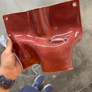 May include: A glossy, dark rose-colored ceramic wall sconce with a sculptural, organic shape. The piece features two circular mounting holes at the top corners and a central opening with a clear electrical cord threaded through the middle.