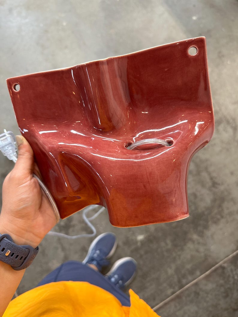 May include: A glossy, dark rose-colored ceramic wall sconce with a sculptural, organic shape. The piece features two circular mounting holes at the top corners and a central opening with a clear electrical cord threaded through the middle.