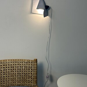 May include: A wall-mounted metal lamp with a folded, sheet-like design that emits a warm glow from behind its curved edge. The fixture features a black socket and a thin, transparent power cord with an inline switch hanging down the wall.