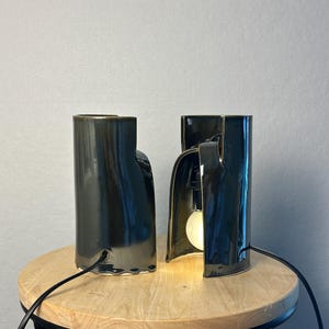 Puede incluir: Dos l&aacute;mparas de mesa de cer&aacute;mica verde oscuro con acabado brillante. Cada l&aacute;mpara presenta una forma cil&iacute;ndrica y escultural con una secci&oacute;n arqueada abierta en la base. Una l&aacute;mpara est&aacute; iluminada por una bombilla redonda, ambas tienen cables negros.