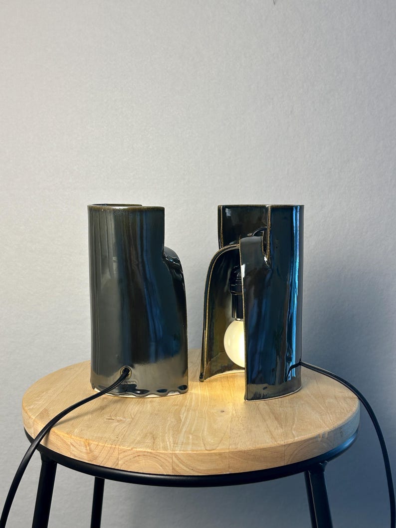 Puede incluir: Dos l&aacute;mparas de mesa de cer&aacute;mica verde oscuro con acabado brillante. Cada l&aacute;mpara presenta una forma cil&iacute;ndrica y escultural con una secci&oacute;n arqueada abierta en la base. Una l&aacute;mpara est&aacute; iluminada por una bombilla redonda, ambas tienen cables negros.