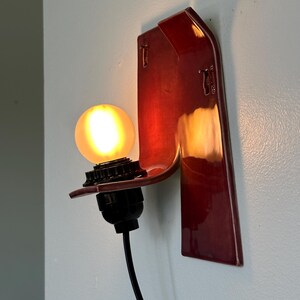 以下が含まれることがあります： 深紅色の光沢仕上げの壁掛け照明器具。器具は、電球ソケットと光る丸い電球を備えた曲線的なデザインです。黒い電源コードがベースから伸びています。器具は水色の壁に取り付けられています。