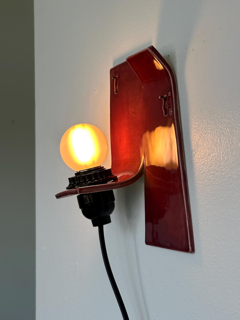 以下が含まれることがあります： 深紅色の光沢仕上げの壁掛け照明器具。器具は、電球ソケットと光る丸い電球を備えた曲線的なデザインです。黒い電源コードがベースから伸びています。器具は水色の壁に取り付けられています。