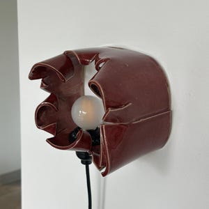 May include: A burgundy ceramic wall sconce with a unique, ruffled design. The sconce features a light bulb and a black electrical cord. The fixture is mounted on a white wall, providing a modern and artistic lighting solution.