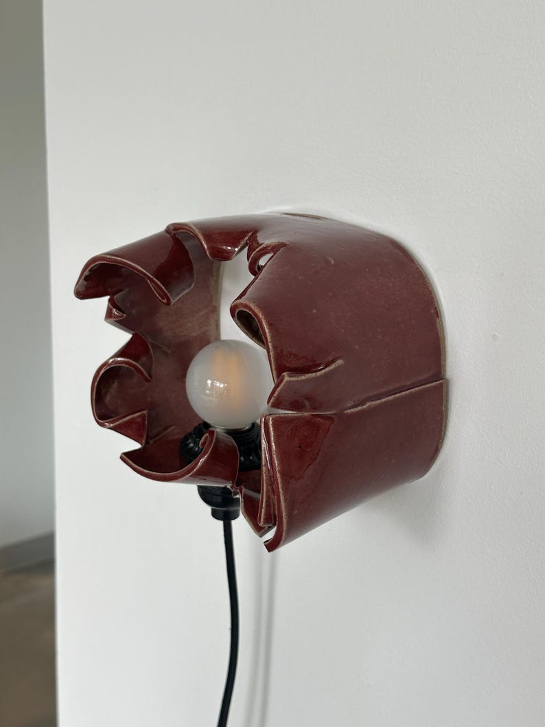 May include: A burgundy ceramic wall sconce with a unique, ruffled design. The sconce features a light bulb and a black electrical cord. The fixture is mounted on a white wall, providing a modern and artistic lighting solution.