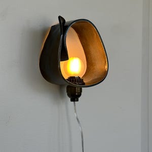 May include: A unique wall sconce with a dark, curved metal shade. The interior is a warm, golden color, and it houses a glowing light bulb. The sconce is mounted on a wall and has a clear power cord.