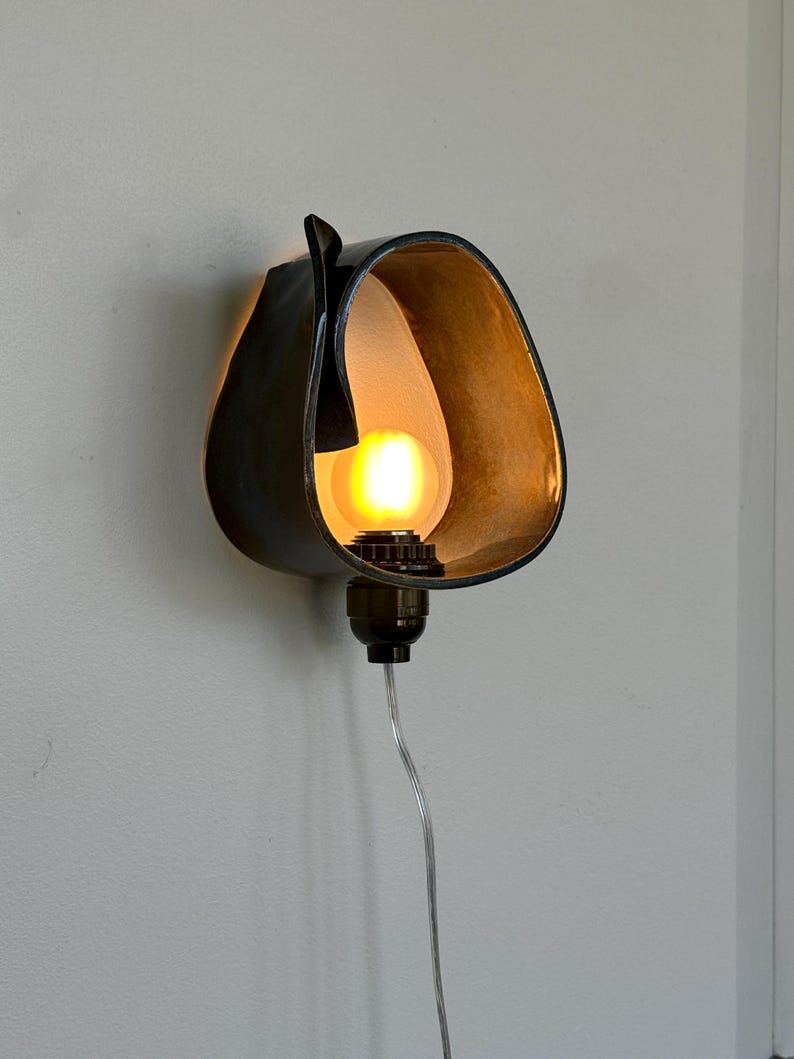 May include: A unique wall sconce with a dark, curved metal shade. The interior is a warm, golden color, and it houses a glowing light bulb. The sconce is mounted on a wall and has a clear power cord.