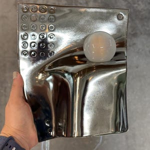 K&ouml;nnte beinhalten: Eine rechteckige Wandleuchte mit reflektierender silberner Chromoberfl&auml;che und wellenf&ouml;rmiger, skulpturaler Struktur. Die Leuchte verf&uuml;gt &uuml;ber ein Raster aus kleinen kreisf&ouml;rmigen Vertiefungen oben links und eine kugelf&ouml;rmige wei&szlig;e Gl&uuml;hbirne.