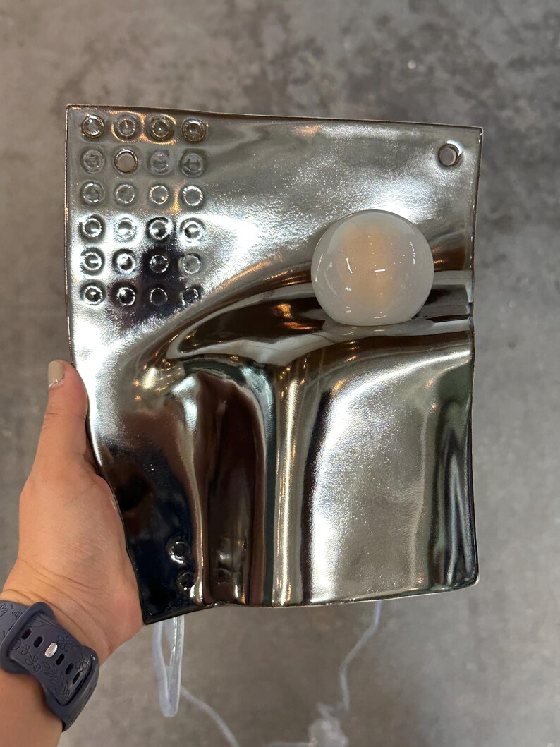 K&ouml;nnte beinhalten: Eine rechteckige Wandleuchte mit reflektierender silberner Chromoberfl&auml;che und wellenf&ouml;rmiger, skulpturaler Struktur. Die Leuchte verf&uuml;gt &uuml;ber ein Raster aus kleinen kreisf&ouml;rmigen Vertiefungen oben links und eine kugelf&ouml;rmige wei&szlig;e Gl&uuml;hbirne.