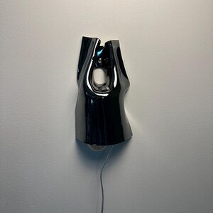 May include: A wall-mounted lamp with a glossy, black, fluid-shaped shade that features a central cutout. The fixture has a reflective surface and includes a thin, light-coloured power cord extending downwards from the base of the lamp structure.