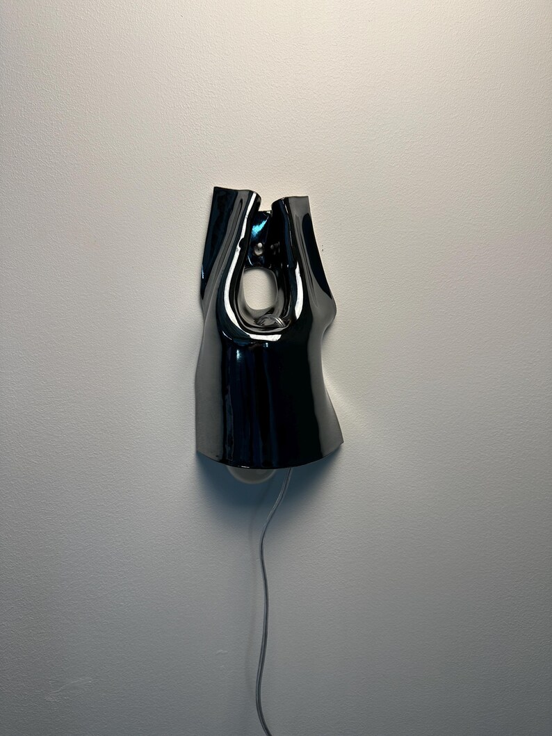 May include: A wall-mounted lamp with a glossy, black, fluid-shaped shade that features a central cutout. The fixture has a reflective surface and includes a thin, light-coloured power cord extending downwards from the base of the lamp structure.