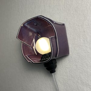 K&ouml;nnte beinhalten: Eine Wandleuchte mit einem gl&auml;nzenden, violetten Keramikschirm in unregelm&auml;&szlig;iger, gefalteter geometrischer Form. Die Leuchte verf&uuml;gt &uuml;ber eine schwarze Fassung mit einer leuchtenden, runden wei&szlig;en Gl&uuml;hbirne und einem transparenten Kabel.