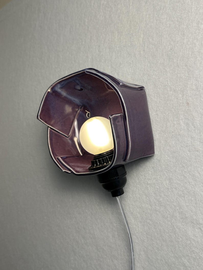 K&ouml;nnte beinhalten: Eine Wandleuchte mit einem gl&auml;nzenden, violetten Keramikschirm in unregelm&auml;&szlig;iger, gefalteter geometrischer Form. Die Leuchte verf&uuml;gt &uuml;ber eine schwarze Fassung mit einer leuchtenden, runden wei&szlig;en Gl&uuml;hbirne und einem transparenten Kabel.