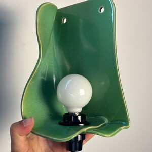 Pu&ograve; includere: Una lampada da parete con un paralume in ceramica verde lucido dalla forma organica, dotato di due fori di montaggio circolari nella parte superiore. Un portalampada nero sostiene una lampadina rotonda bianca opaca e un cavo nero esce dalla base.