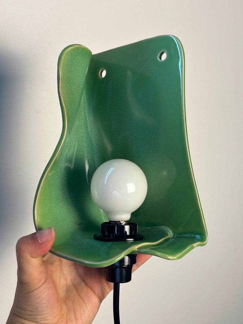 Pu&ograve; includere: Una lampada da parete con un paralume in ceramica verde lucido dalla forma organica, dotato di due fori di montaggio circolari nella parte superiore. Un portalampada nero sostiene una lampadina rotonda bianca opaca e un cavo nero esce dalla base.
