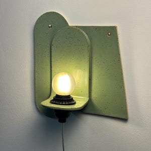 May include: A wall-mounted light fixture featuring a light green ceramic backplate with dark speckles. The fixture has an arched shape and a small shelf holding a round, frosted glass light bulb. A black socket base connects to a thin power cord.