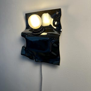May include: A wall-mounted lamp featuring a glossy black, irregularly shaped ceramic backplate with a wavy, folded texture. A single round, frosted white light bulb is mounted on the upper portion of the plate, emitting a warm glow.