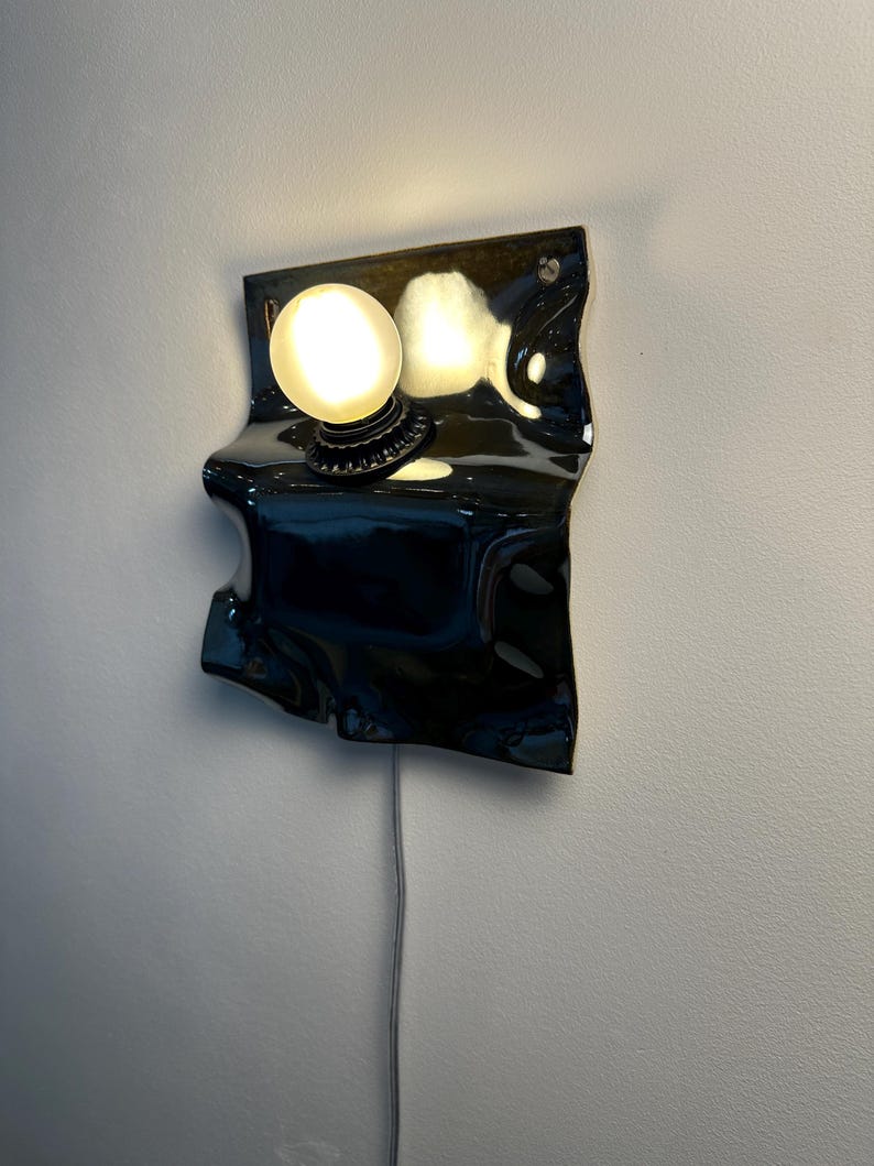 May include: A wall-mounted lamp featuring a glossy black, irregularly shaped ceramic backplate with a wavy, folded texture. A single round, frosted white light bulb is mounted on the upper portion of the plate, emitting a warm glow.