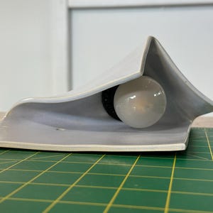May include: A light gray ceramic wall sconce with a folded, angular silhouette. The fixture features a central circular opening housing a single round white light bulb. The ceramic material has a smooth, matte finish and a distinct geometric shape.