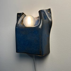 May include: A wall-mounted ceramic sconce featuring a deep blue, glossy glaze with an organic, asymmetrical shape. A single, round, frosted white light bulb is nestled within the upper curve of the fixture, emitting a soft, warm glow.