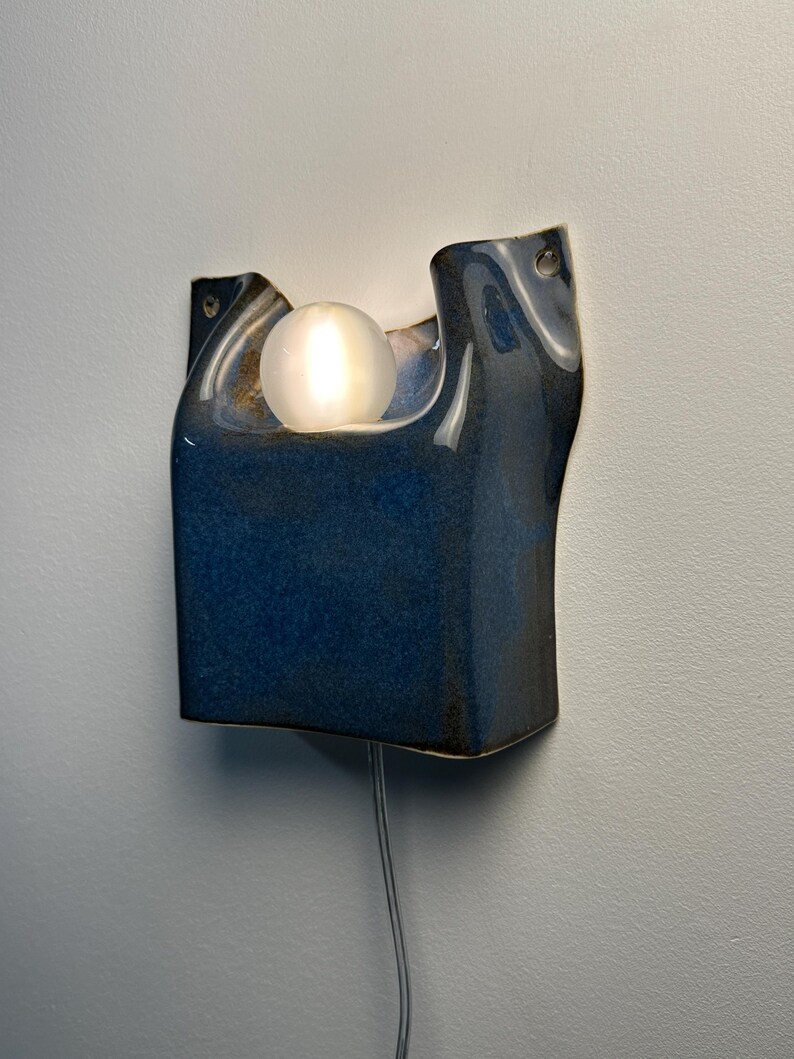 May include: A wall-mounted ceramic sconce featuring a deep blue, glossy glaze with an organic, asymmetrical shape. A single, round, frosted white light bulb is nestled within the upper curve of the fixture, emitting a soft, warm glow.
