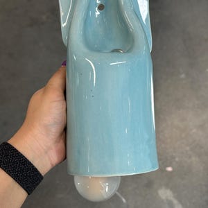 May include: A light blue ceramic wall sconce with a smooth, glossy finish. The fixture features a vertical, elongated body with a curved top section containing a mounting hole and a circular light bulb protruding from the bottom opening.