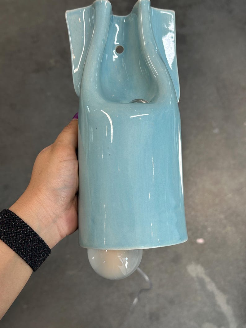 May include: A light blue ceramic wall sconce with a smooth, glossy finish. The fixture features a vertical, elongated body with a curved top section containing a mounting hole and a circular light bulb protruding from the bottom opening.
