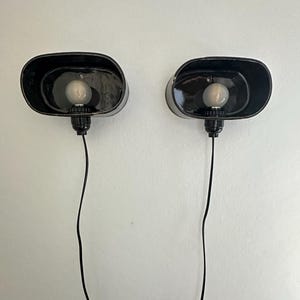 May include: Two black, oval wall sconces with visible light bulbs. Each sconce has a black cord and is mounted on a white wall. The sconces have a minimalist design, with a simple, industrial aesthetic.